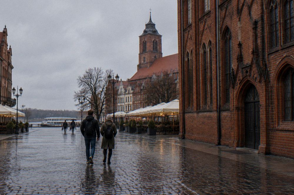 Two visitors seen from behind strolling across the cobbles of Toruń's Old Town beside a red-brick Gothic facade, cafe umbrellas at the edge.