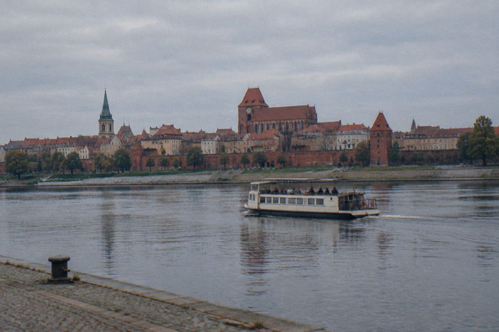 A small plain unmarked tour boat on the wide Vistula with the red-brick Toruń skyline behind, calm water, a few passengers small on deck.