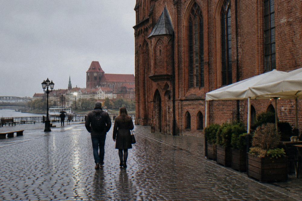 Two visitors seen from behind strolling across the cobbles of Toruń's Old Town beside a red-brick Gothic facade, cafe umbrellas at the edge.