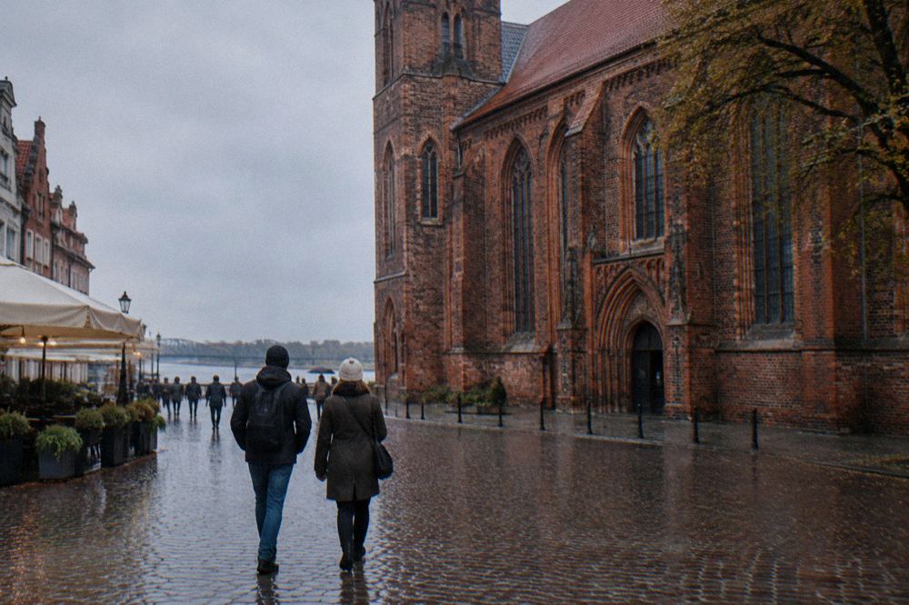 Two visitors seen from behind strolling across the cobbles of Toruń's Old Town beside a red-brick Gothic facade, cafe umbrellas at the edge.