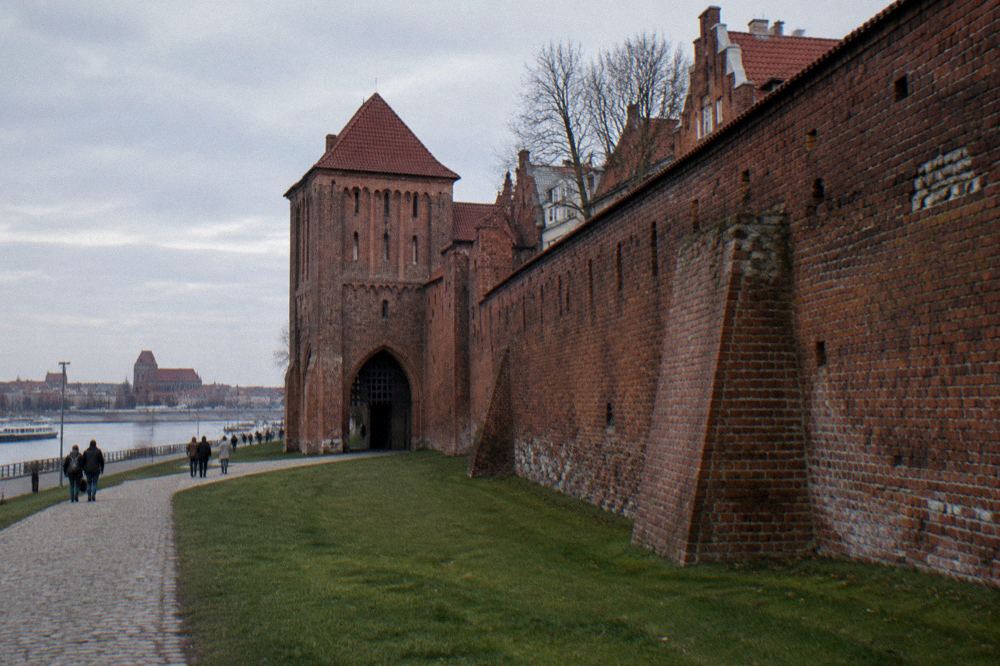A long stretch of weathered red-brick medieval town wall with a fortified gate, grass at its foot, long shadows, Toruń.