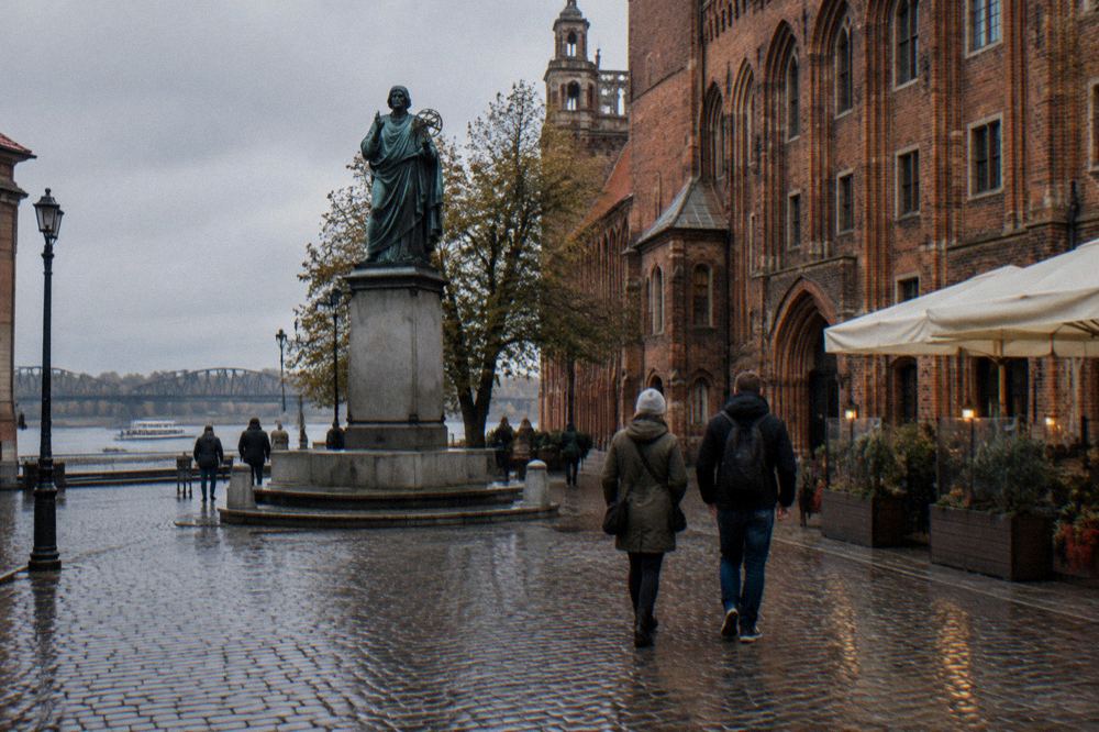 Two visitors seen from behind strolling across the cobbles of Toruń's Old Town beside a red-brick Gothic facade, cafe umbrellas at the edge.