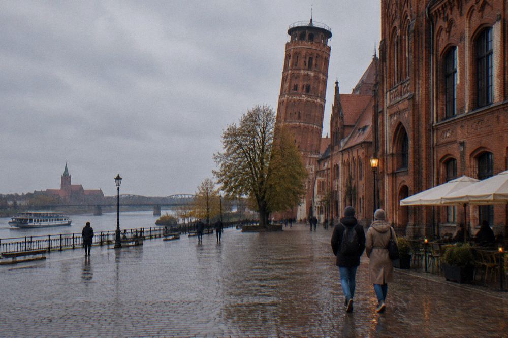 Two visitors seen from behind strolling across the cobbles of Toruń's Old Town beside a red-brick Gothic facade, cafe umbrellas at the edge.