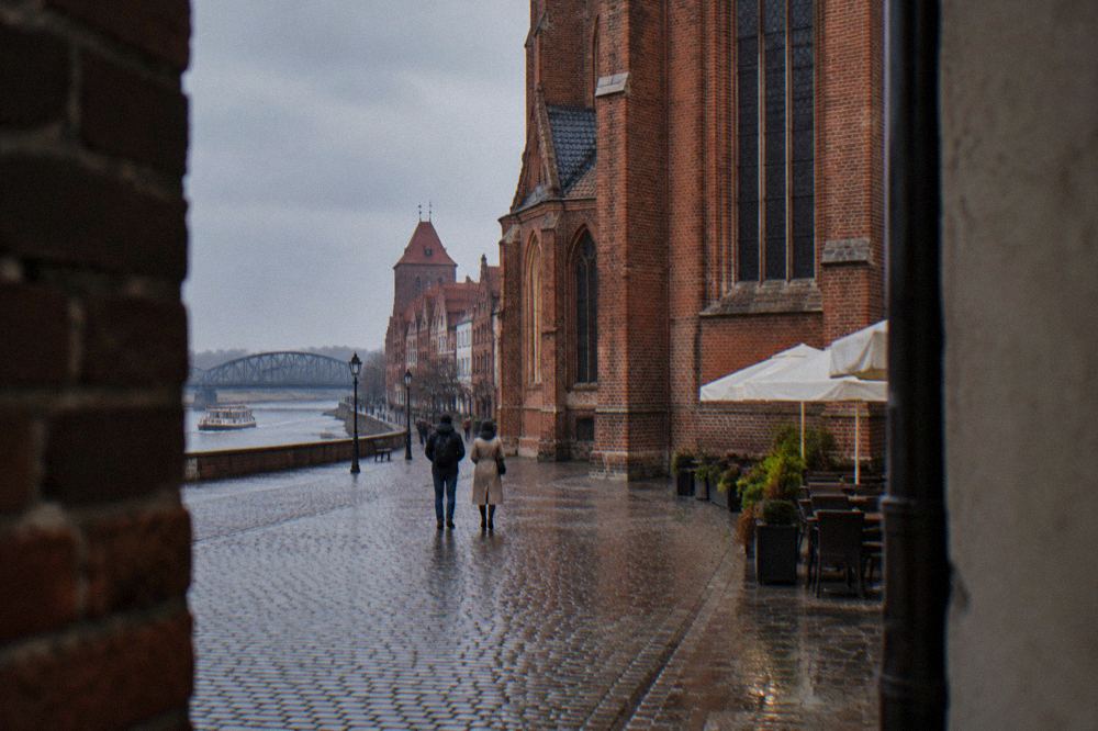 Two visitors seen from behind strolling across the cobbles of Toruń's Old Town beside a red-brick Gothic facade, cafe umbrellas at the edge.