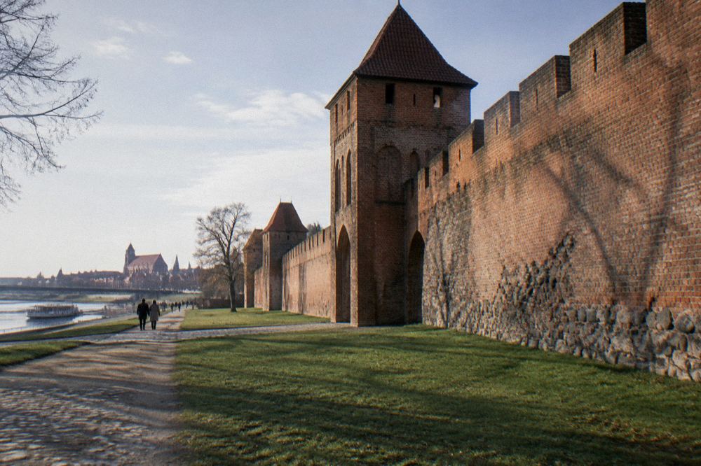 A long stretch of weathered red-brick medieval town wall with a fortified gate, grass at its foot, long shadows, Toruń.
