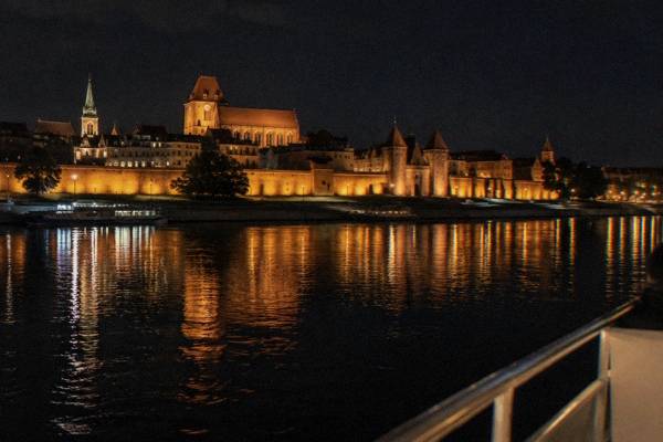 The floodlit red-brick walls and gates of Toruń reflected in the black Vistula, seen from a boat deck at night, the rail of the boat in the corner, Philadelphia Boulevard
