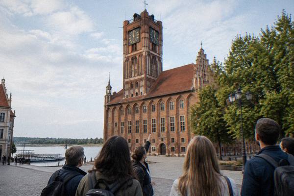 A small group with a guide seen from behind gathered by the red-brick Old Town hall in Toruń, the guide pointing up at the tower, Old Town Hall and Tower