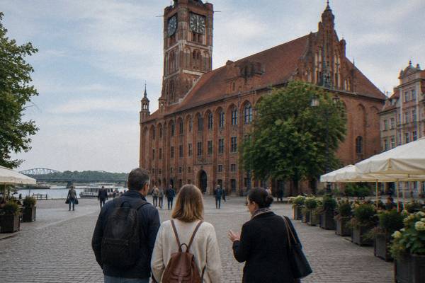 Two visitors with a private guide strolling past the Old Town hall in Toruń, a walking pace, seen from behind, cafe umbrellas at the edge, Old Town Market Square