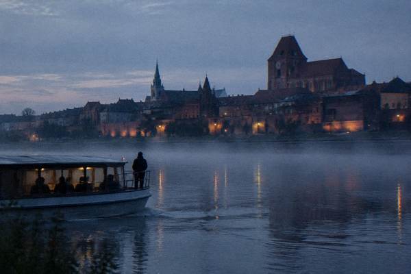 A small tour boat with a guide silhouetted at the rail drifting through low river mist on the Vistula at blue hour, the Toruń walls dark and lit by a few lamps, Kepa Bazarowa Viewpoint