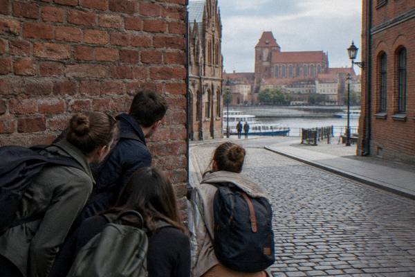 A small group of friends crouching and peering around the corner of an old brick lane in Toruń as if hunting for clues, seen from behind, cobbles, Szeroka Street