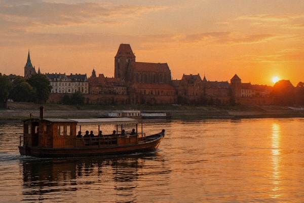 A traditional wooden river boat gliding on the Vistula at sunset, the brick Old Town skyline glowing orange behind it, a few silhouetted passengers, Philadelphia Boulevard
