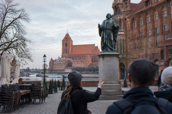 A guide talking to a few people from behind in front of the bronze astronomer statue in Toruń's Old Town square, empty cafe chairs stacked, Nicolaus Copernicus Monument