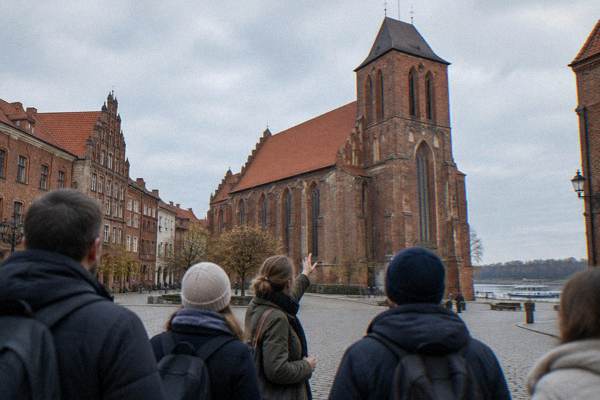 A local guide gesturing toward a tall Gothic brick church while a few visitors listen, all seen from behind, Old Town square in Toruń, St Mary's Church