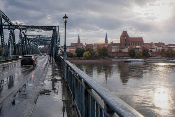The long steel bridge over the Vistula with the red-brick Toruń skyline across the water after a rain shower, a dark car crossing, puddles on the road, Pilsudski Bridge