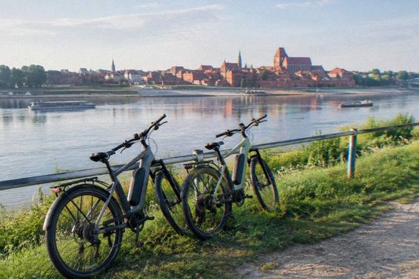 Two electric bikes leaning on a railing on the grassy Vistula levee path in Toruń, the brick skyline small across the river, Kepa Bazarowa Viewpoint