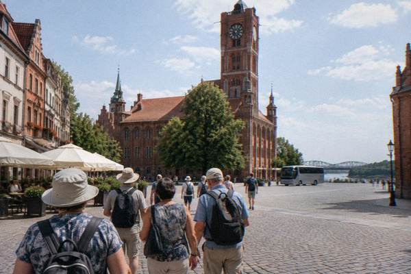 A tour group seen from behind arriving in the sunny Old Town market square in Toruń, the town hall tower ahead, a coach stopped far off without markings