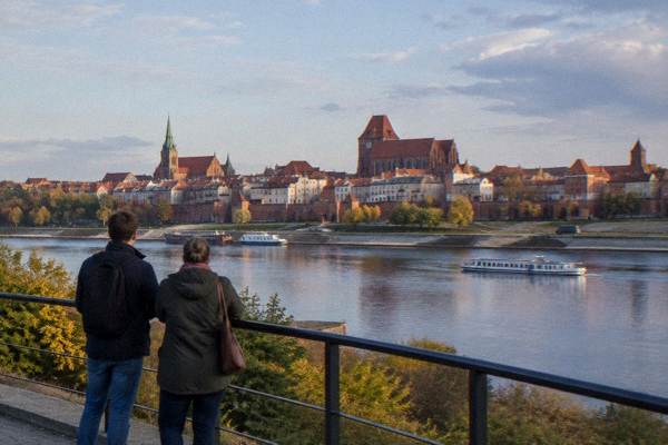 A visitor and a driver-guide from behind standing at a viewpoint rail looking over the Vistula to the brick Toruń skyline, a dark minivan parked far off without markings, Philadelphia Boulevard