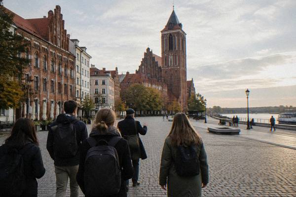 A group of friends from behind walking across the quiet New Town square in Toruń with a guide, tall church tower beyond, New Town Market Square