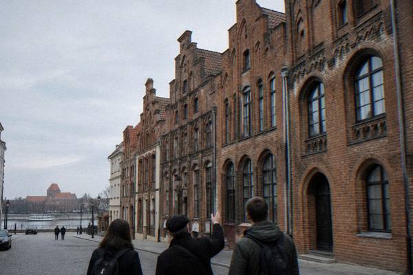 A guide and two visitors from behind stopping at the Gothic brick townhouses on Kopernika street in Toruń, gables high above, Copernicus House