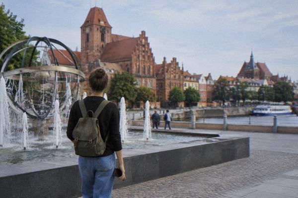 A solo traveller seen from behind pausing by a modern fountain near the Old Town in Toruń, holding a phone low at their side, brick gables ahead, Cosmopolis Fountain