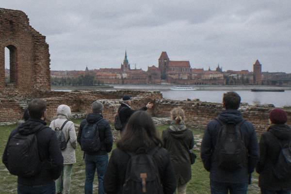 A group on the grass among low brick castle ruins in Toruń, listening to a guide, grey sky, the Old Town roofline beyond, all seen from behind, Teutonic Castle Ruins