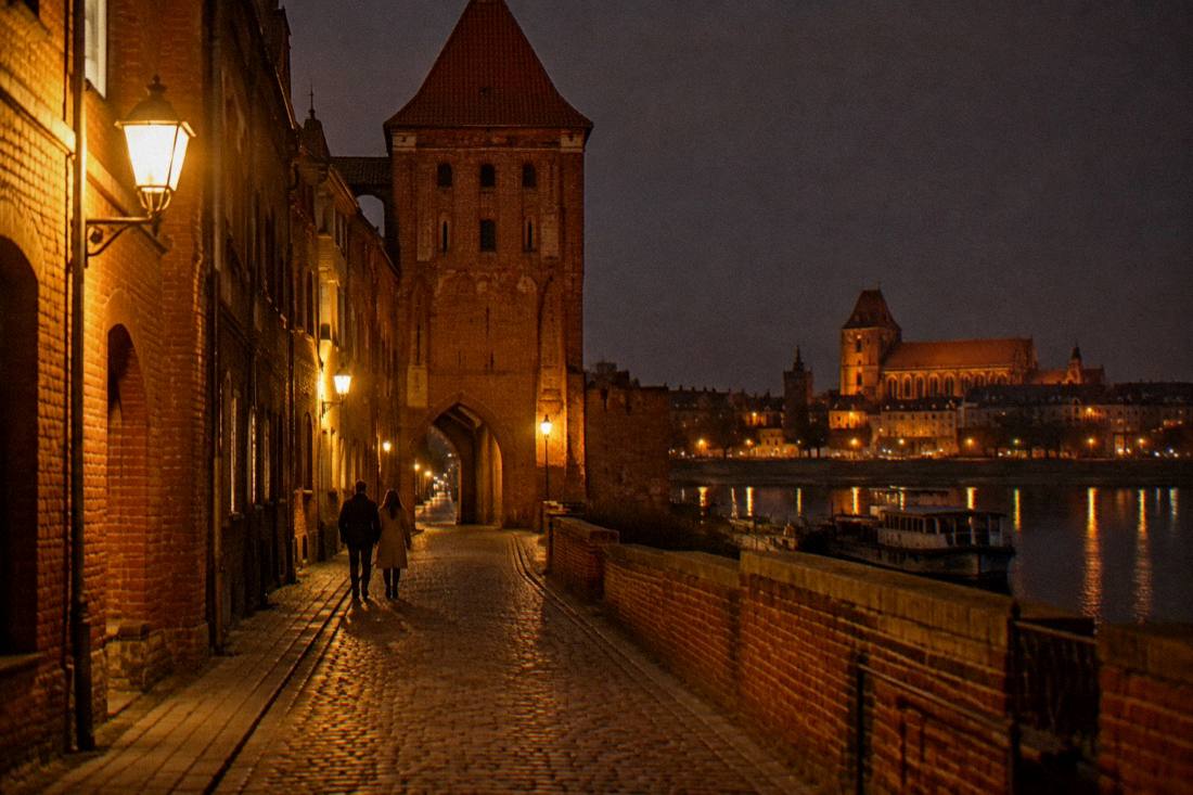 A cobbled lane in Toruń's Old Town at night with warm lamps on brick walls