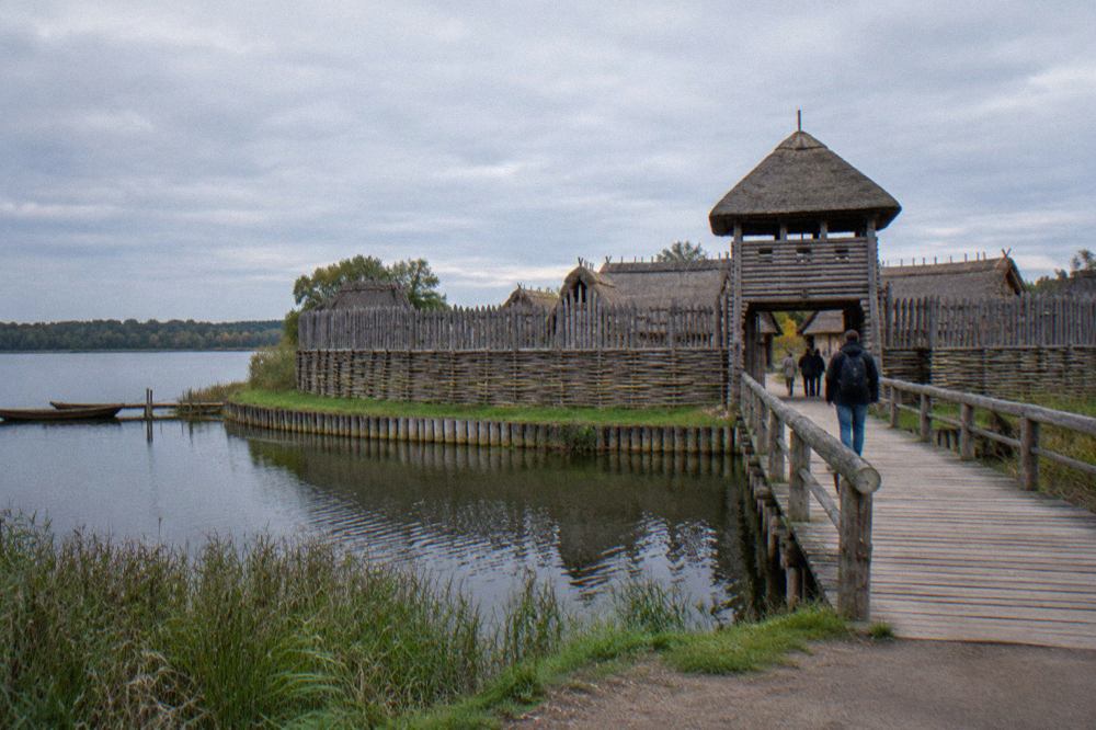 Reconstructed wooden ramparts and gate of an archaeological settlement beside a lake, with a footbridge leading to the entrance