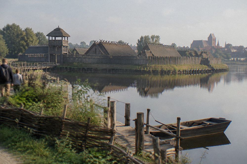 Timber palisade walls and thatched longhouse roofs of a reconstructed prehistoric village seen across still water