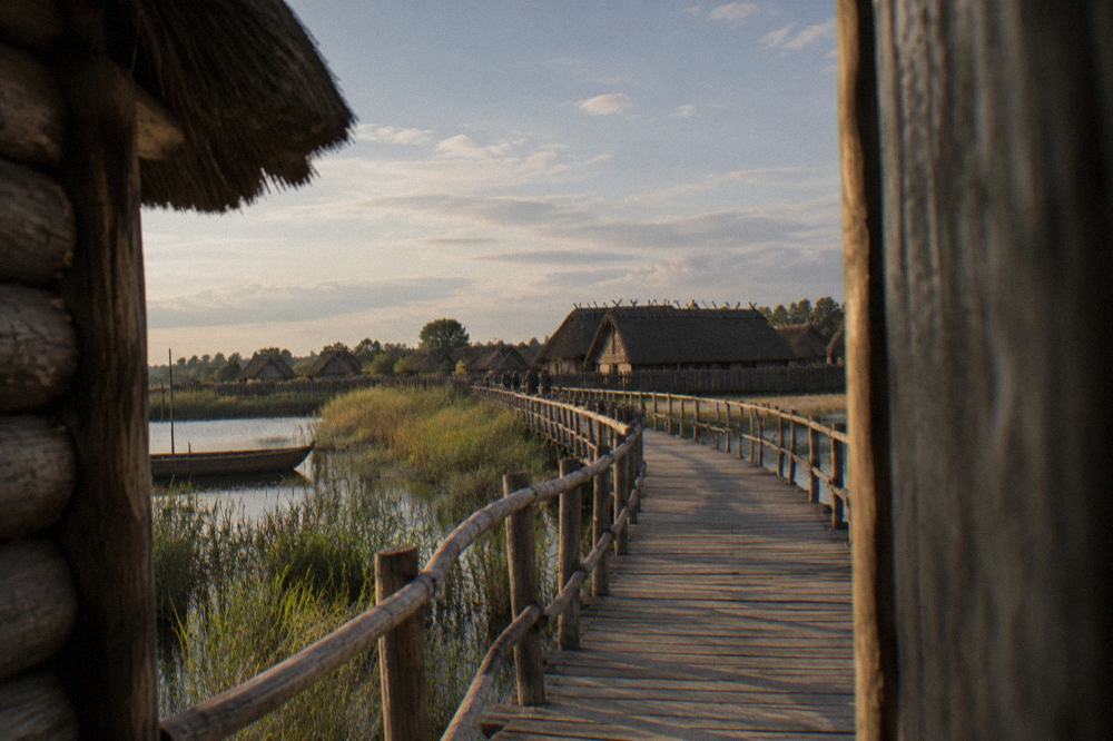 Wooden walkway crossing wetland toward reconstructed prehistoric houses under a wide afternoon sky
