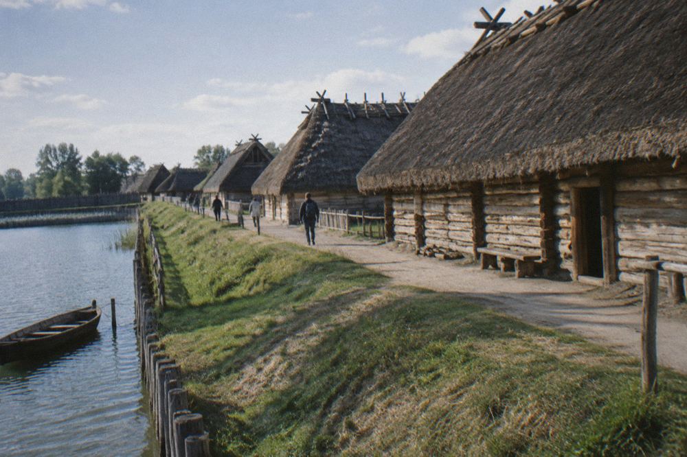 Row of reconstructed timber longhouses with thatched roofs along an earthen embankment at the archaeological park