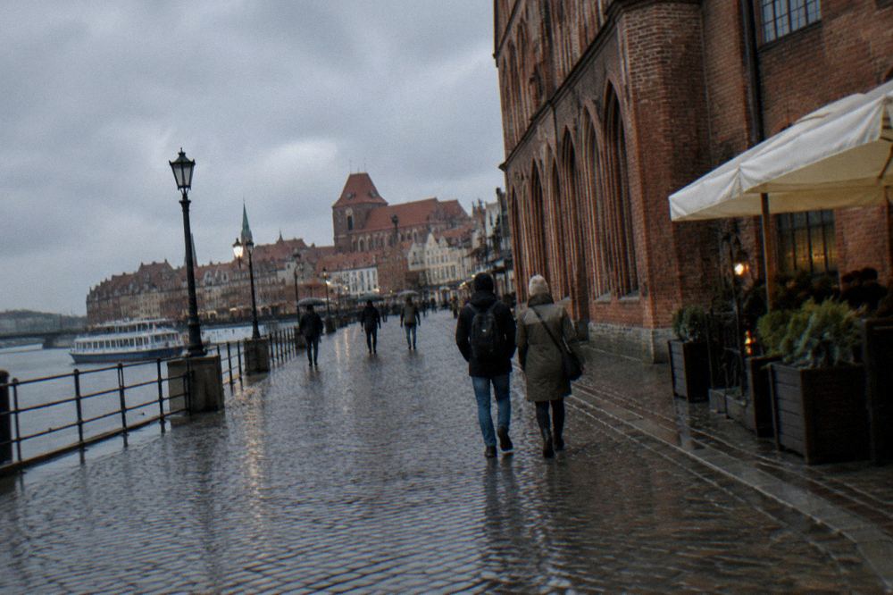 Two visitors seen from behind strolling across the cobbles of Toruń's Old Town beside a red-brick Gothic facade, cafe umbrellas at the edge.