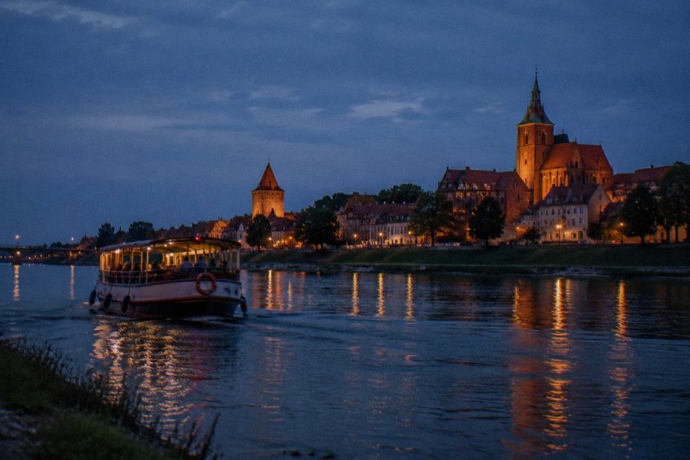 A boat on the Vistula river at dusk with the lit towers of a medieval old town on the far bank