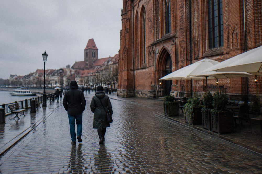 Two visitors seen from behind strolling across the cobbles of Toruń's Old Town beside a red-brick Gothic facade, cafe umbrellas at the edge.