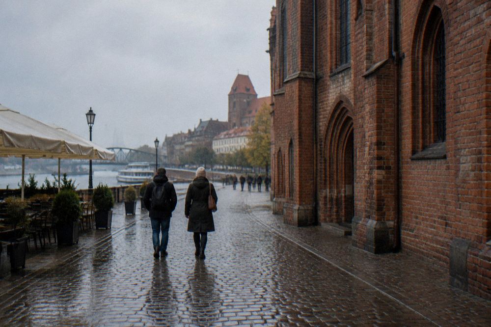 Two visitors seen from behind strolling across the cobbles of Toruń's Old Town beside a red-brick Gothic facade, cafe umbrellas at the edge.