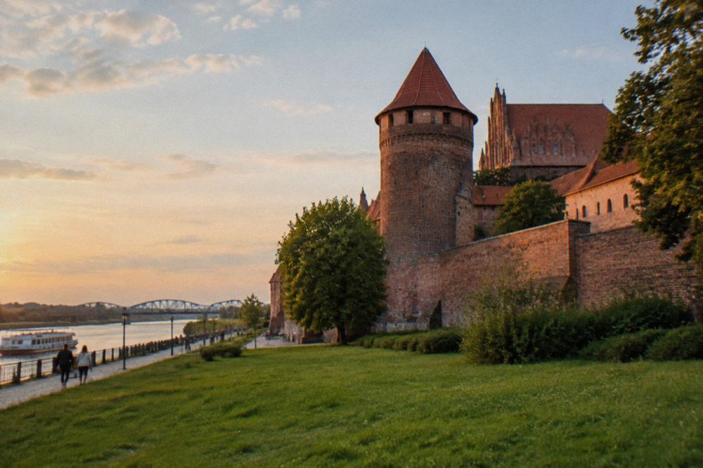 A red-brick Teutonic castle with a cylindrical tower seen from a green lawn