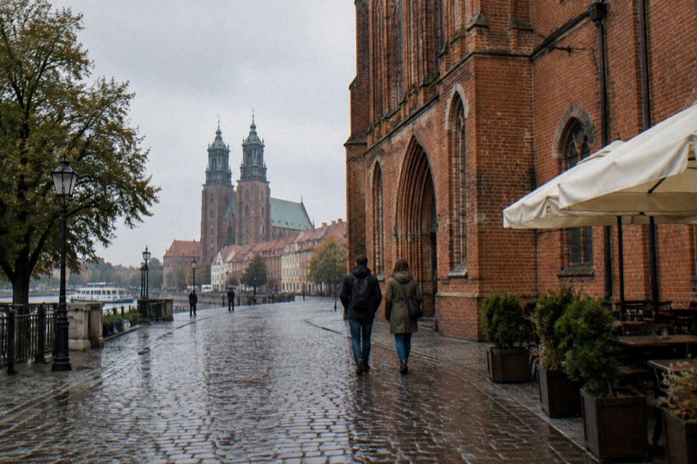 Two visitors seen from behind strolling across the cobbles of Toruń's Old Town beside a red-brick Gothic facade, cafe umbrellas at the edge.