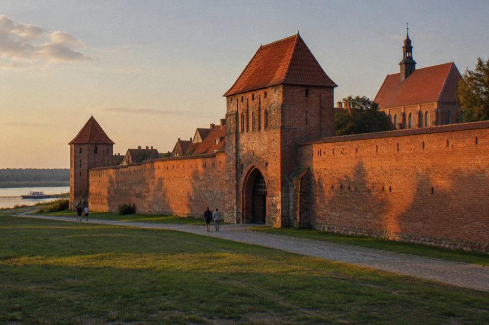 A long stretch of weathered red-brick medieval town wall with a fortified gate, grass at its foot, long shadows, Toruń.
