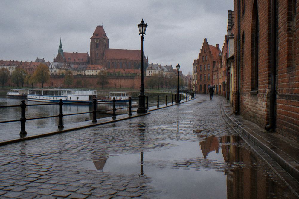 A rain-washed cobbled lane in Toruń's Old Town, wet brick gables reflected in puddles, one person with an umbrella far off.