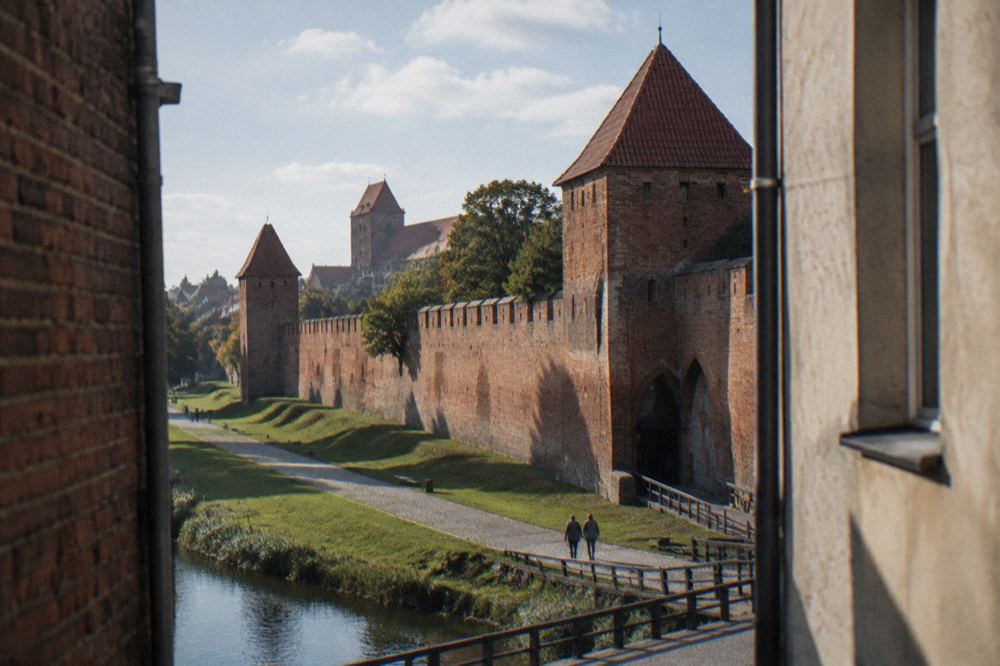 A long stretch of weathered red-brick medieval town wall with a fortified gate, grass at its foot, long shadows, Toruń.