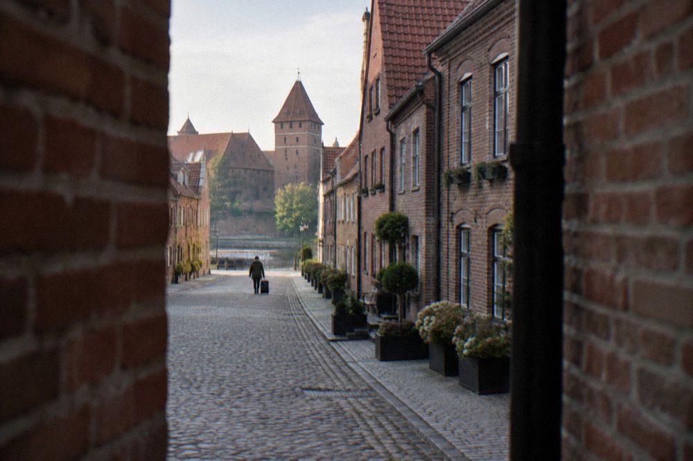 A calm Old Town street in Malborkwith brick townhouses converted to small hotels, cobbles, planters at the doors, a suitcase being wheeled far off.