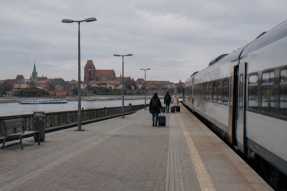A plain unmarked regional train standing at an empty station platform in Płock, a few travellers with suitcases small and from behind.