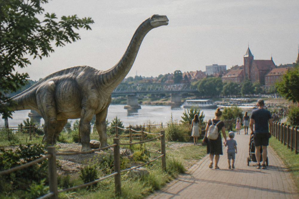 Families walking a paved path beside a large model dinosaur at an outdoor park