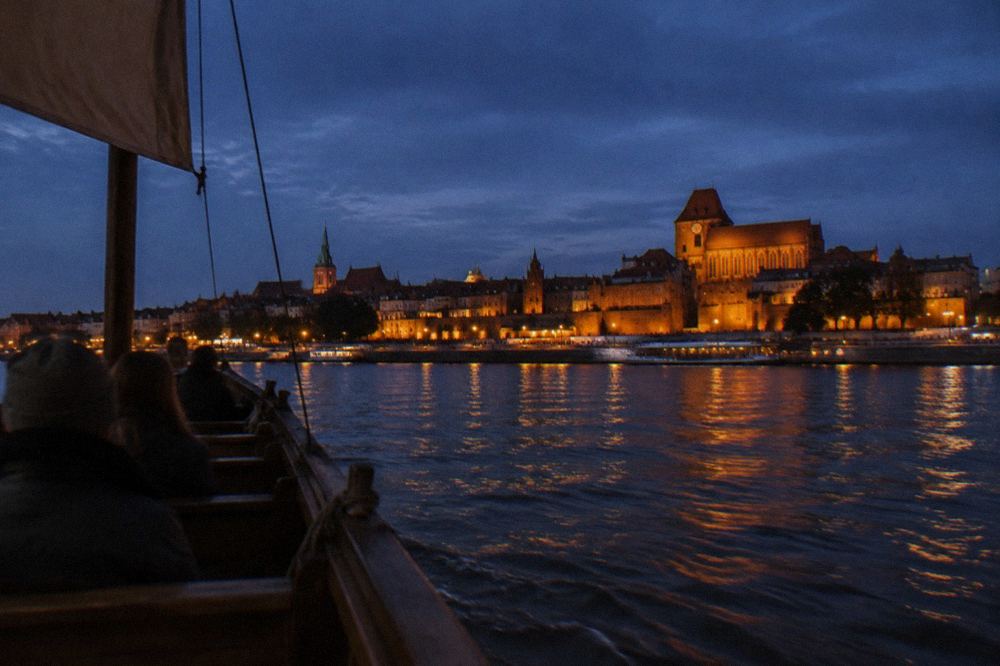 Traditional wooden boat crossing the Vistula past the illuminated Toruń waterfront at night