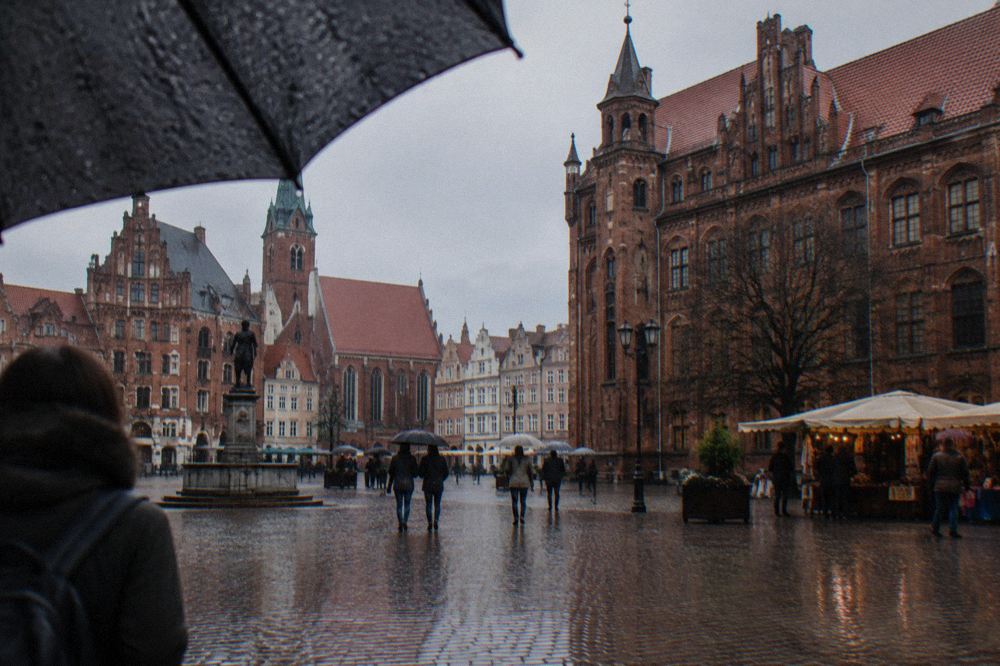 Cobblestone square in the Toruń Old Town under an umbrella in light rain, with gothic brick facades behind