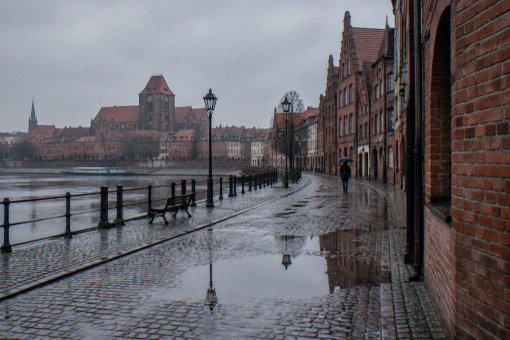 A rain-washed cobbled lane in Toruń's Old Town, wet brick gables reflected in puddles, one person with an umbrella far off.