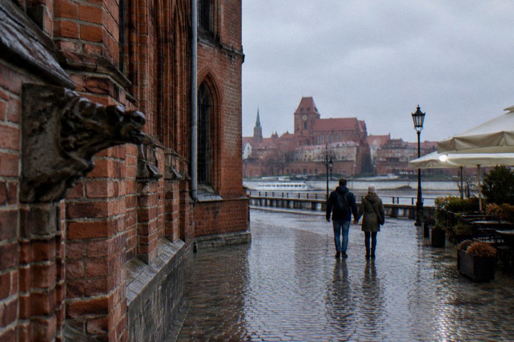 Two visitors seen from behind strolling across the cobbles of Toruń's Old Town beside a red-brick Gothic facade, cafe umbrellas at the edge.