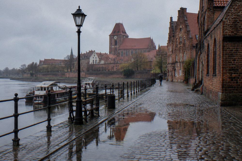 A rain-washed cobbled lane in Toruń's Old Town, wet brick gables reflected in puddles, one person with an umbrella far off.