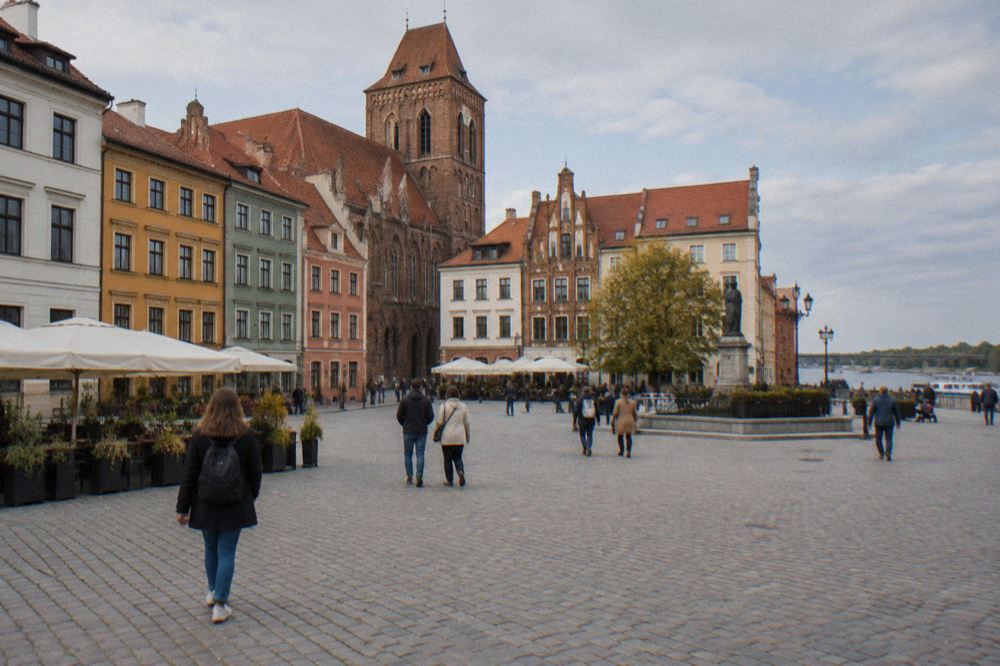 Pedestrians crossing a cobbled square bounded by colourful townhouses in Toruń's Old Town