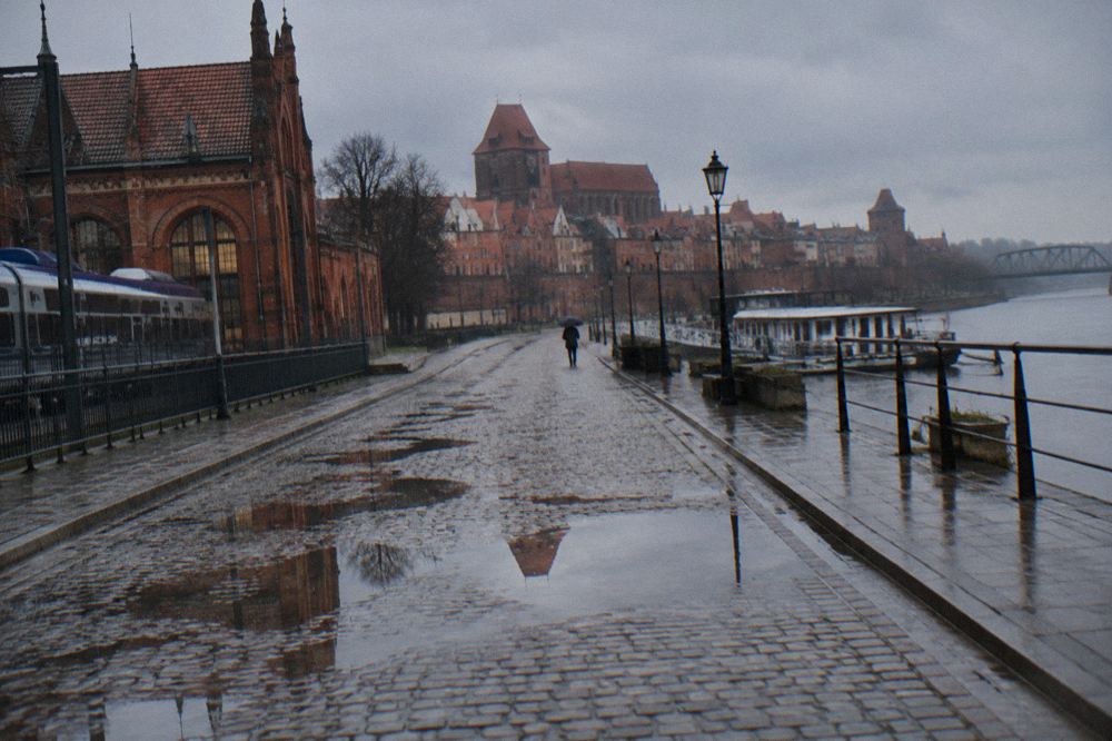 A rain-washed cobbled lane in Toruń's Old Town, wet brick gables reflected in puddles, one person with an umbrella far off.
