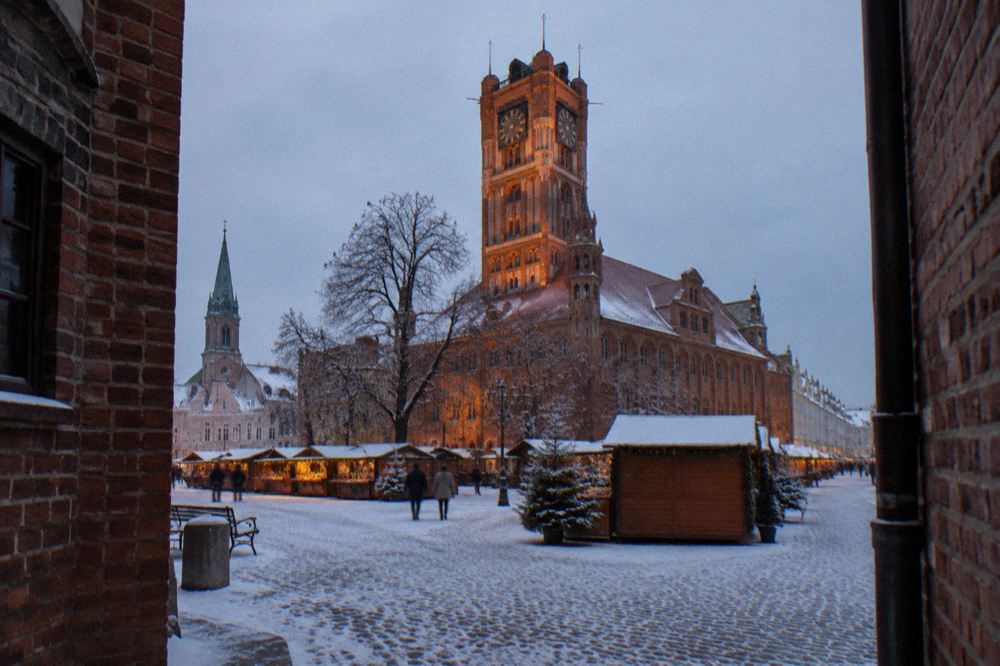 Snow-covered market square in Toruń with the Old Town Hall tower lit at dusk and wooden market stalls